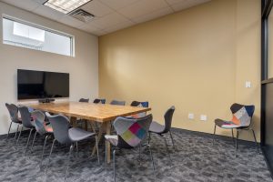 A large conference room with a beautiful wooden conference table, many chairs, and a large tv equipped with a webcam.