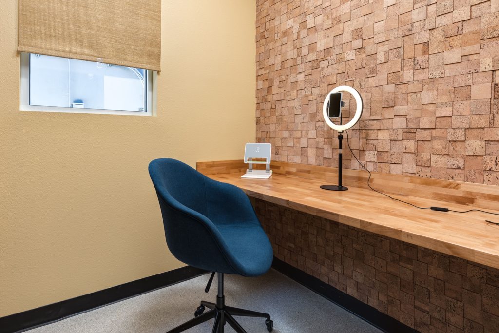 A small office with one cream wall and one brick wall. A ring light and laptop stand are on the wooden desktop.
