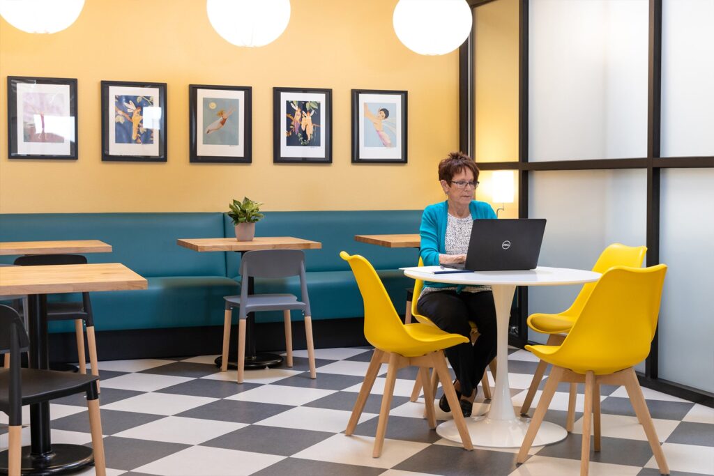 A woman sits in the cafe working on her laptop at one of many small tables.